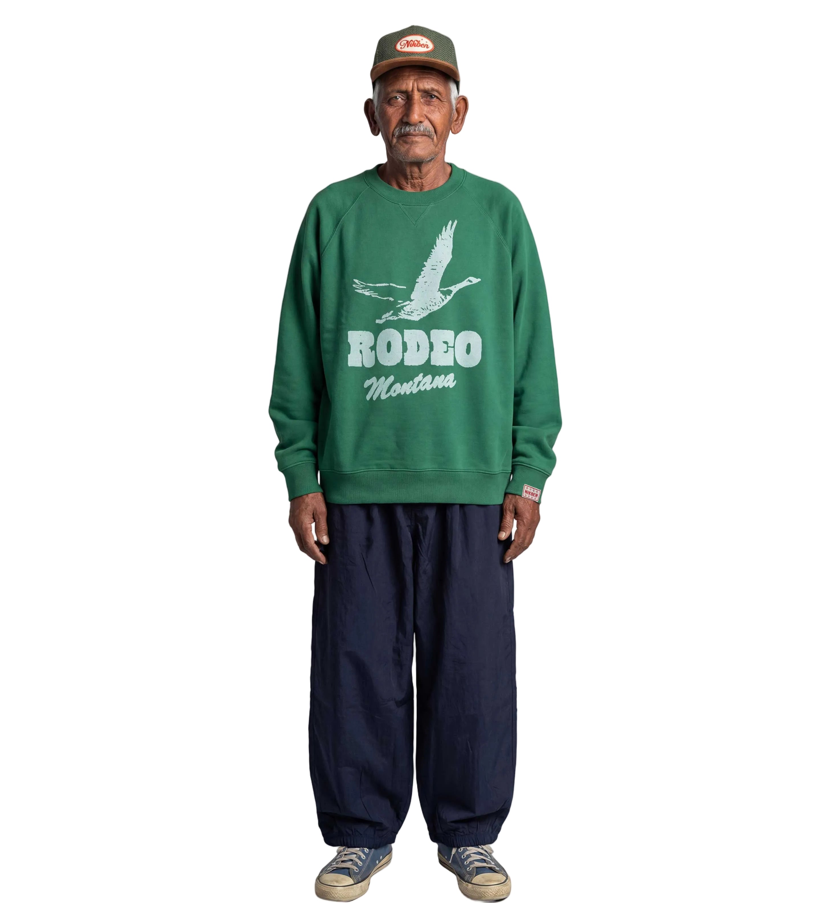 An older man stands against a light gray background wearing a green crewneck sweatshirt with a white 'RODEO Montana' graphic, dark navy pants, and a green cap.