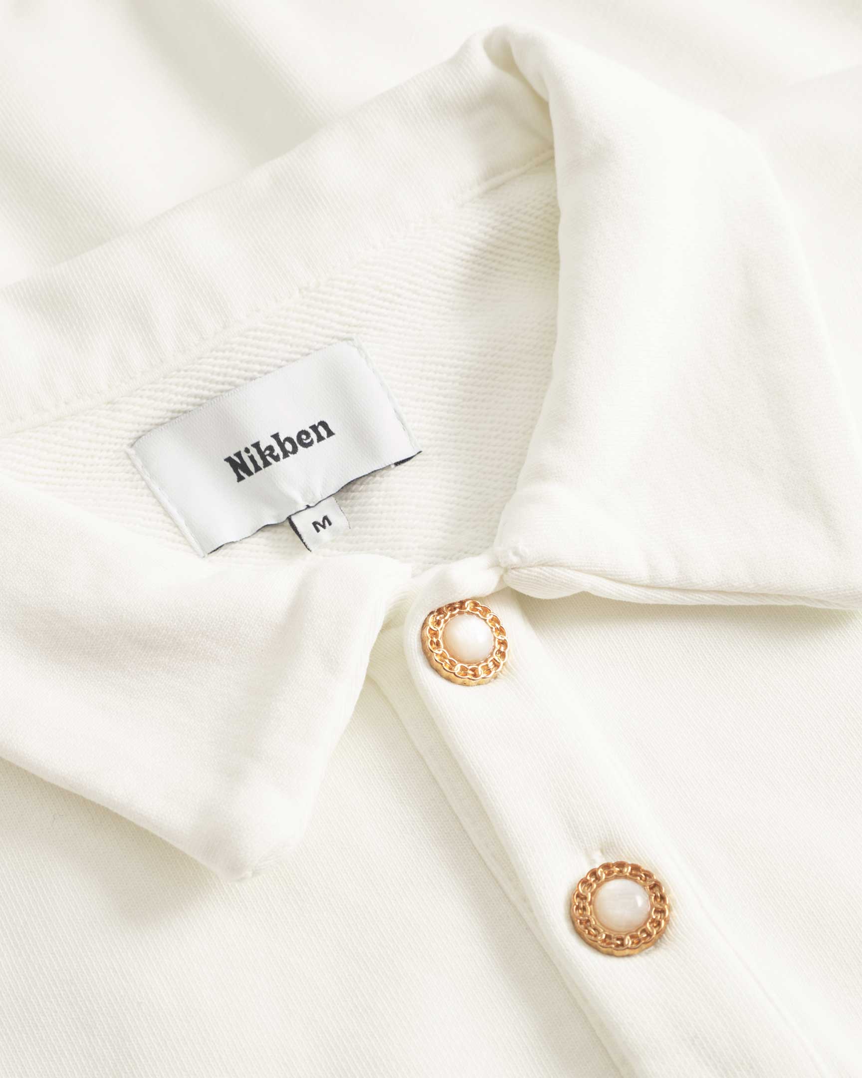 Close up of collar and button closure on a white and green long sleeved polo shirt whit embroidered NB logo and ribbed cuffs