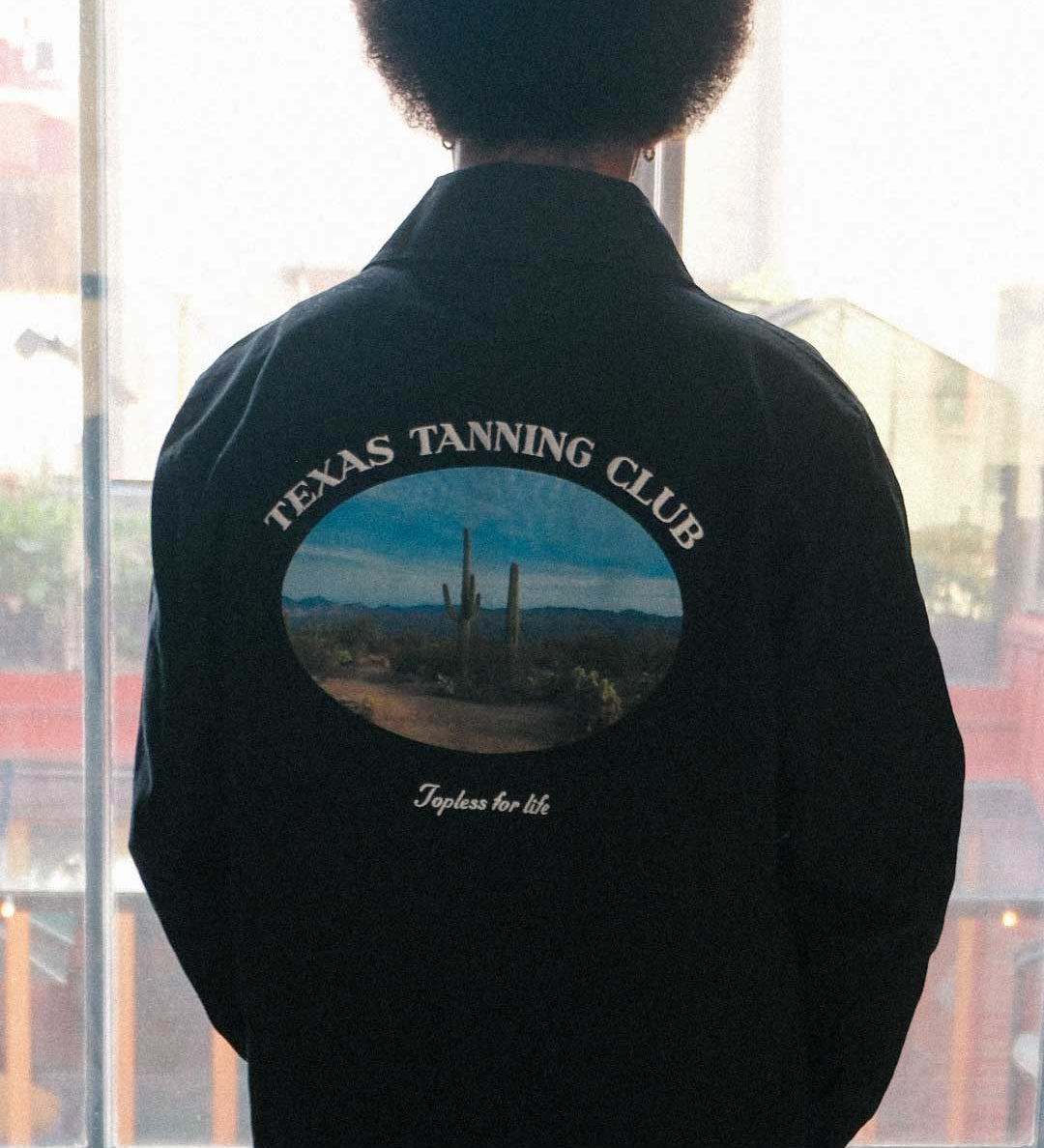model wearing a black overshirt with 'Texas Tanning Club' logo and desert scene on the back, 