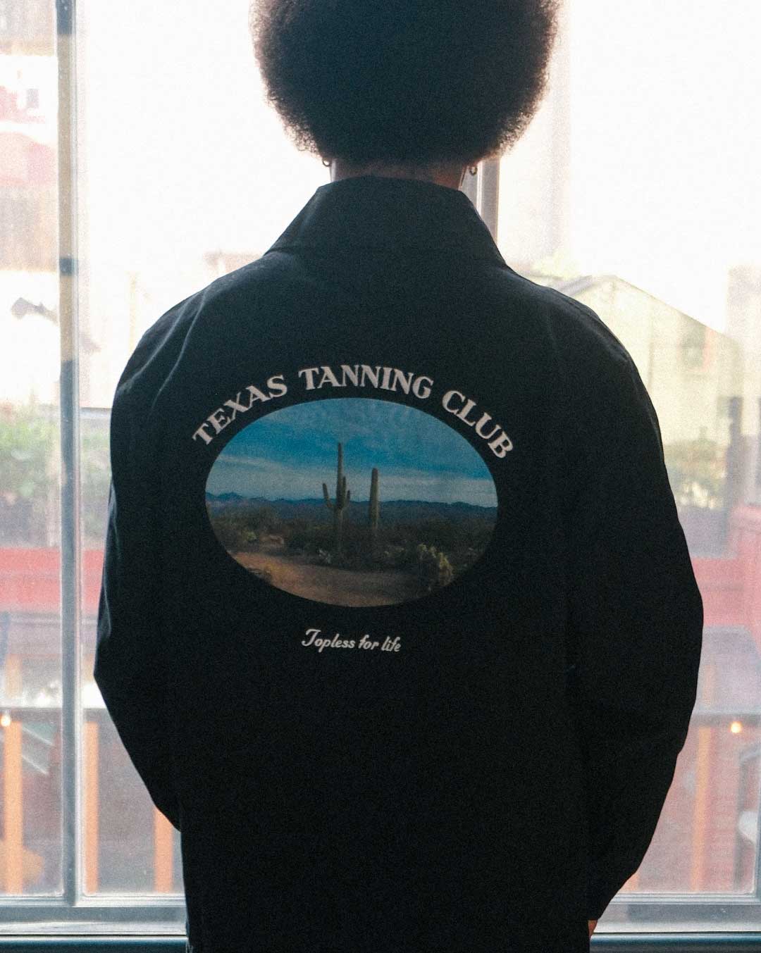 model wearing a black overshirt with 'Texas Tanning Club' logo and desert scene on the back, 