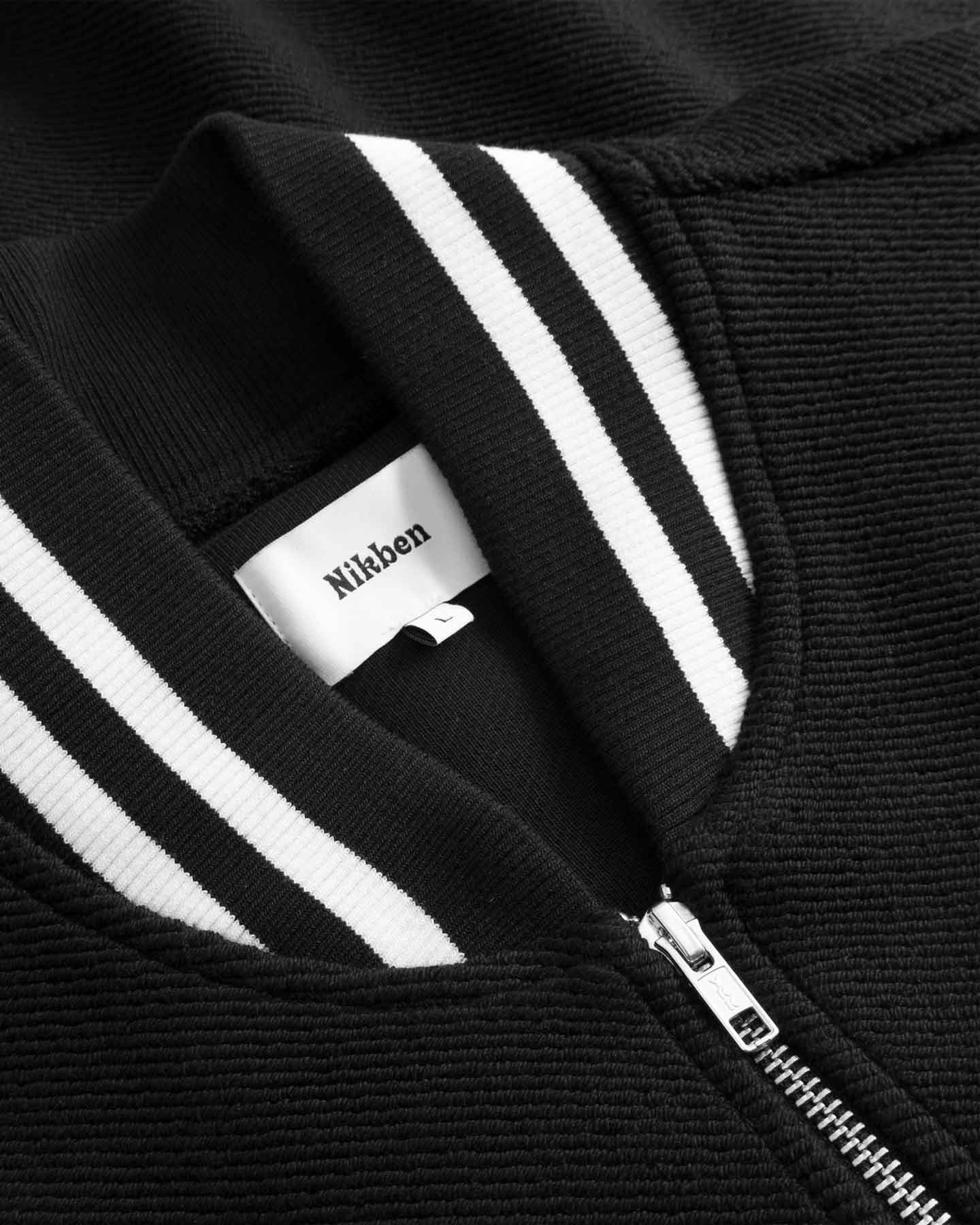 Close-up of black bomber jacket with white stripes and a visible brand label.