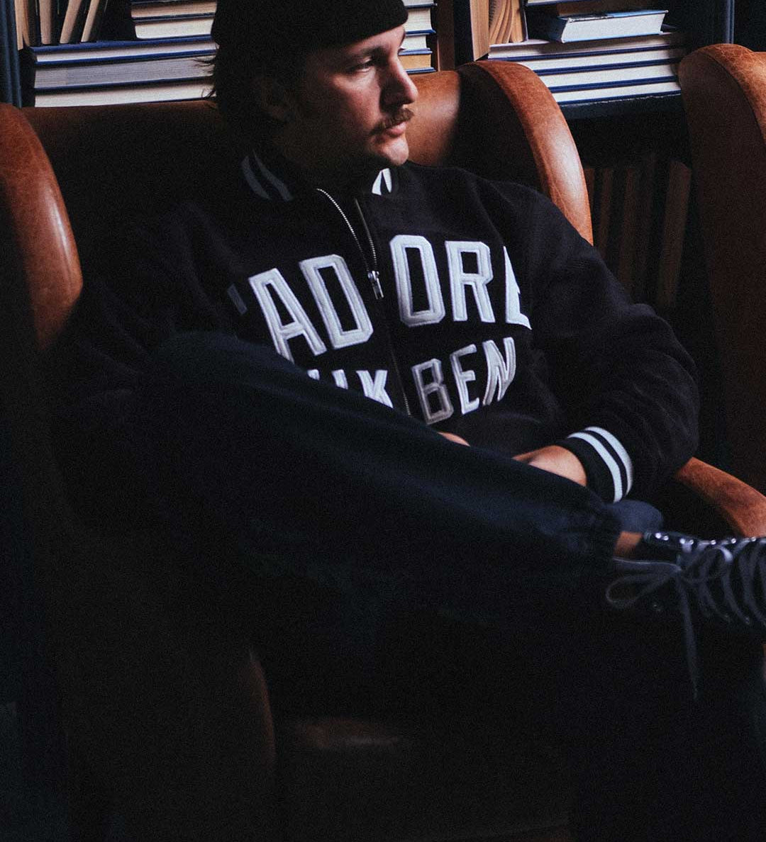 Model wearing a black bomber jacket with white text sitting on a chair