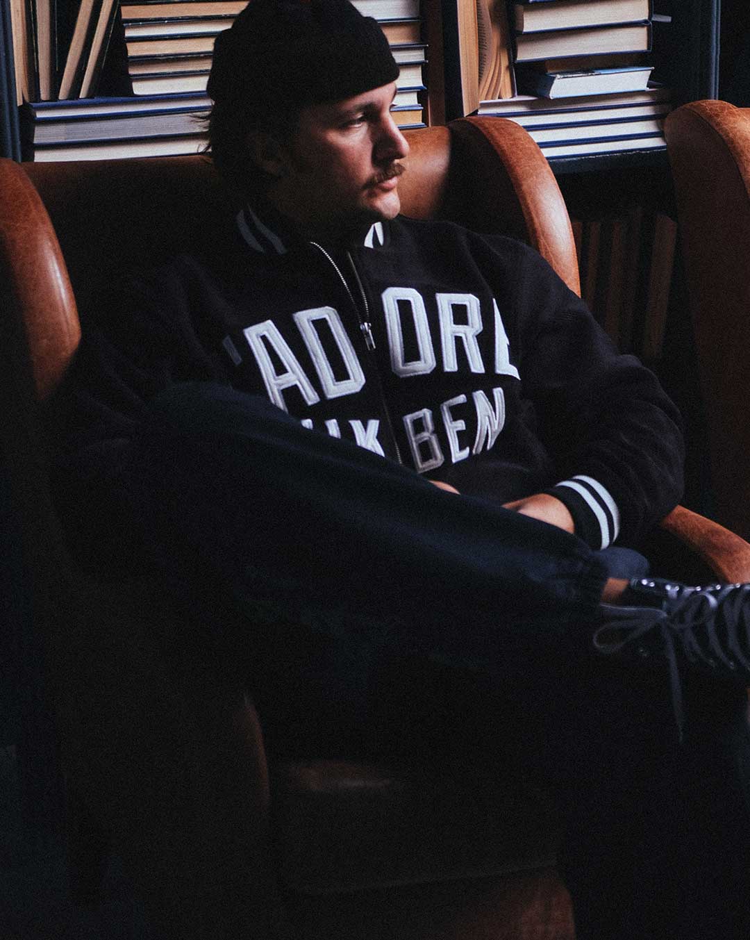 Model wearing a black bomber jacket with white text sitting on a chair