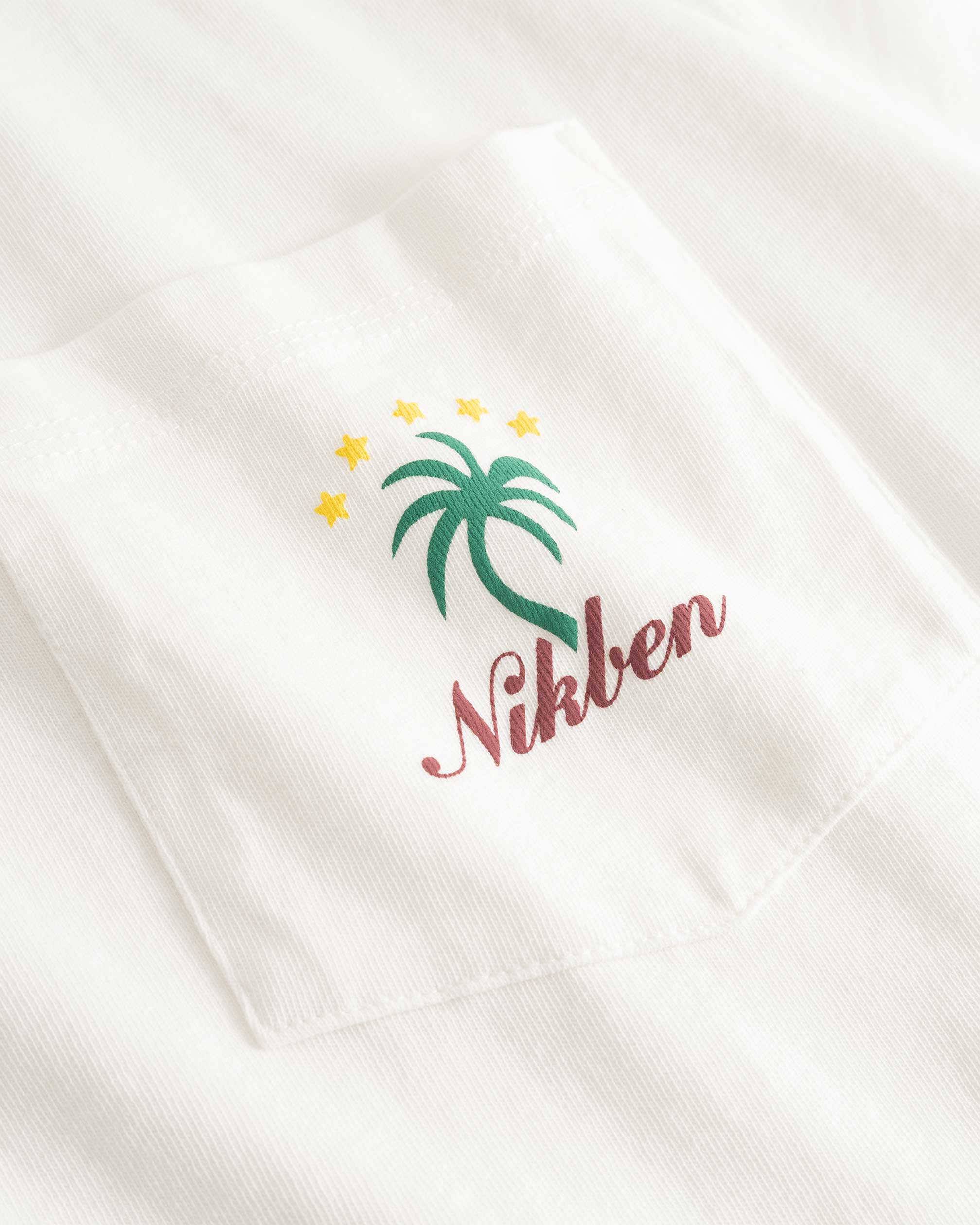 Closeup view of green palm print and red NIKBEN logo on front pocket of white t-shirt