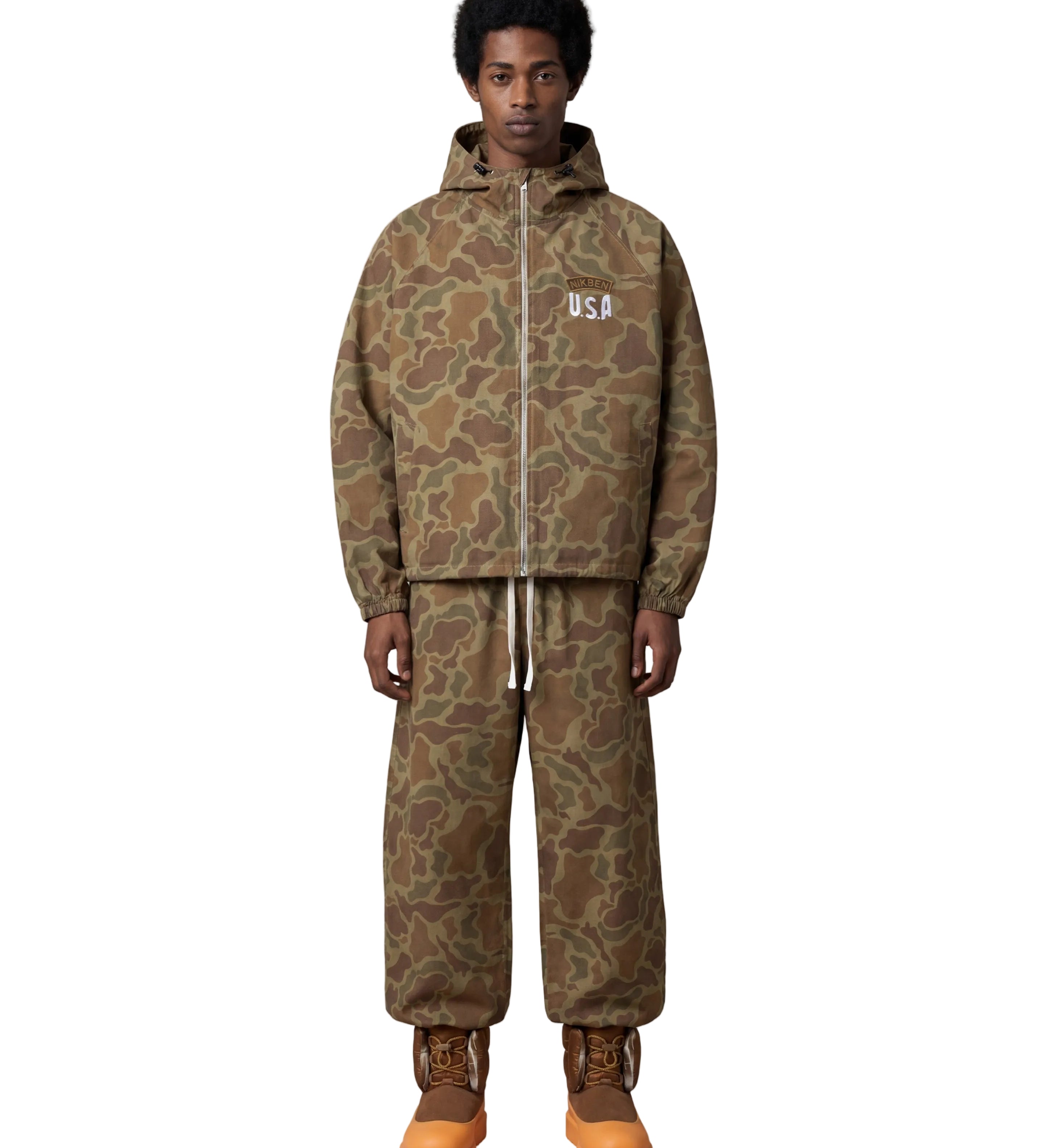 A model stands against a light gray background wearing a matching camouflage hooded jacket and sweatpants set, with brown and orange work boots. The jacket has a white 'USA' logo on the chest.