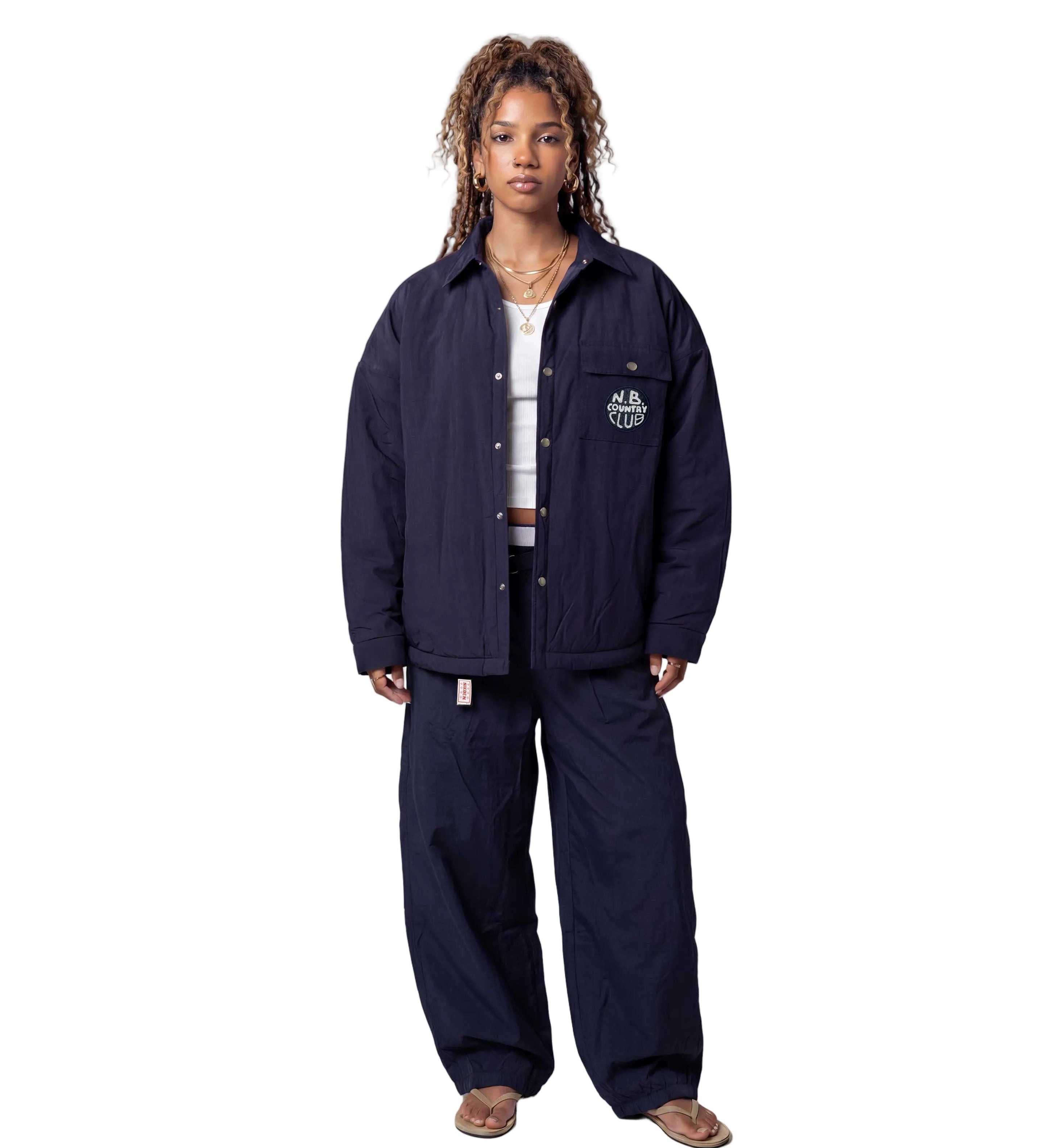 A model stands against a light gray studio background wearing a dark navy blue quilted jacket and matching wide-leg pants, styled with a white t-shirt,
