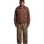 An elderly man stands against a light gray background wearing a brown and rust-orange striped zip-up fleece jacket, brown camouflage pants, a brown cap, and blue sneakers.