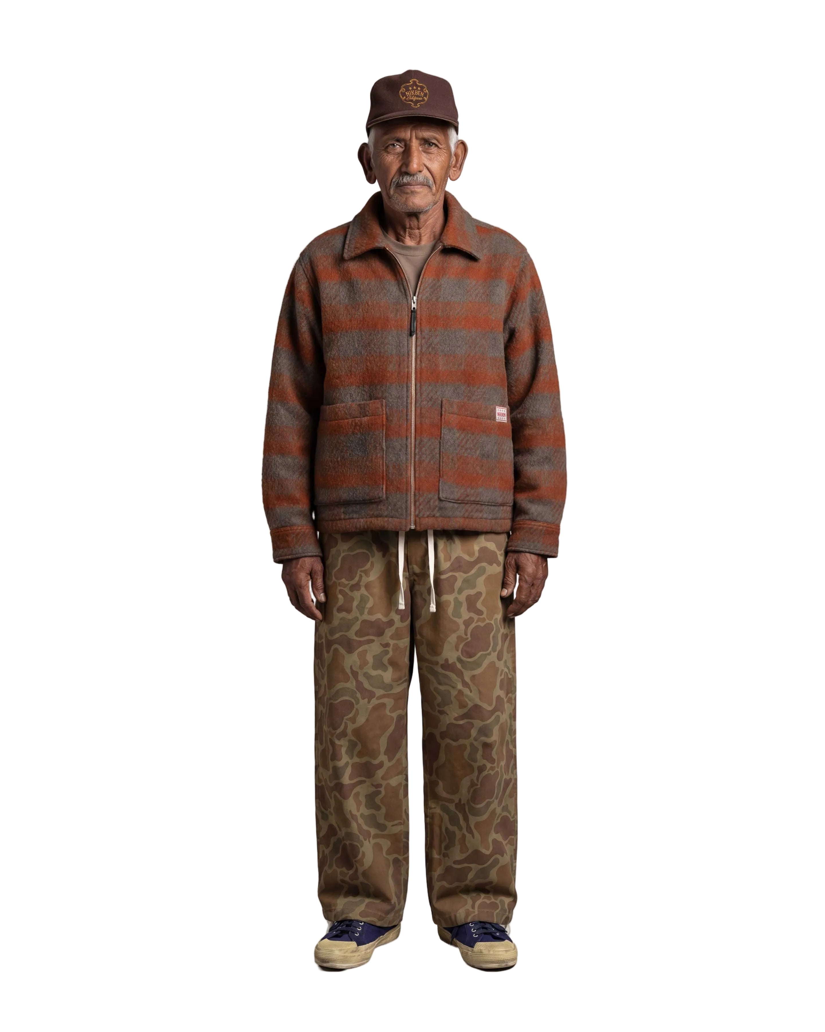 An elderly man stands against a light gray background wearing a brown and rust-orange striped zip-up fleece jacket, brown camouflage pants, a brown cap, and blue sneakers.