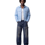 A model stands against a light gray studio background wearing a light blue denim jacket over a white t-shirt, dark blue faded jeans, and off-white sneakers.