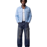 A model stands against a light gray studio background wearing a light blue denim jacket over a white t-shirt, dark blue faded jeans, and off-white sneakers.