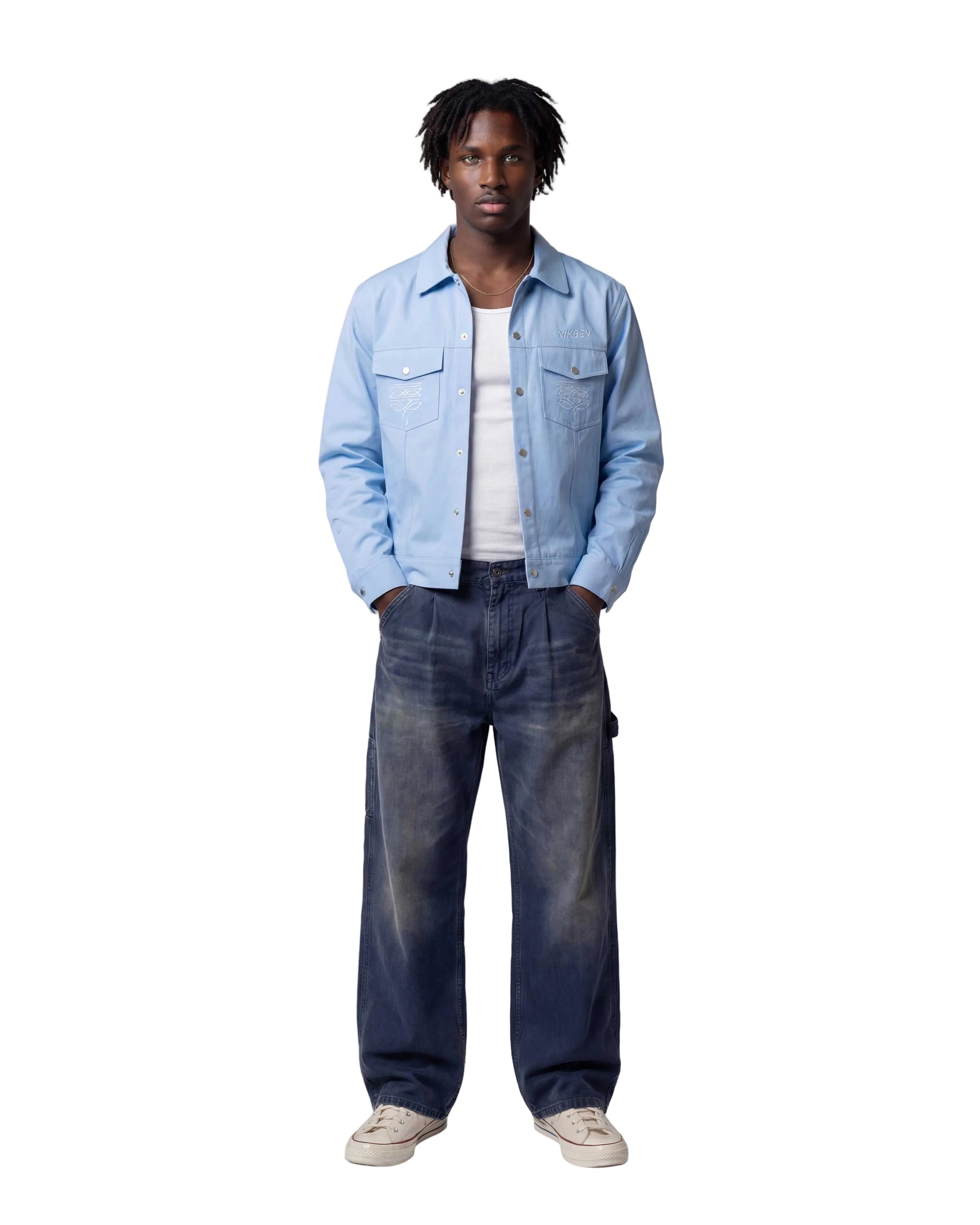 A model stands against a light gray studio background wearing a light blue denim jacket over a white t-shirt, dark blue faded jeans, and off-white sneakers.