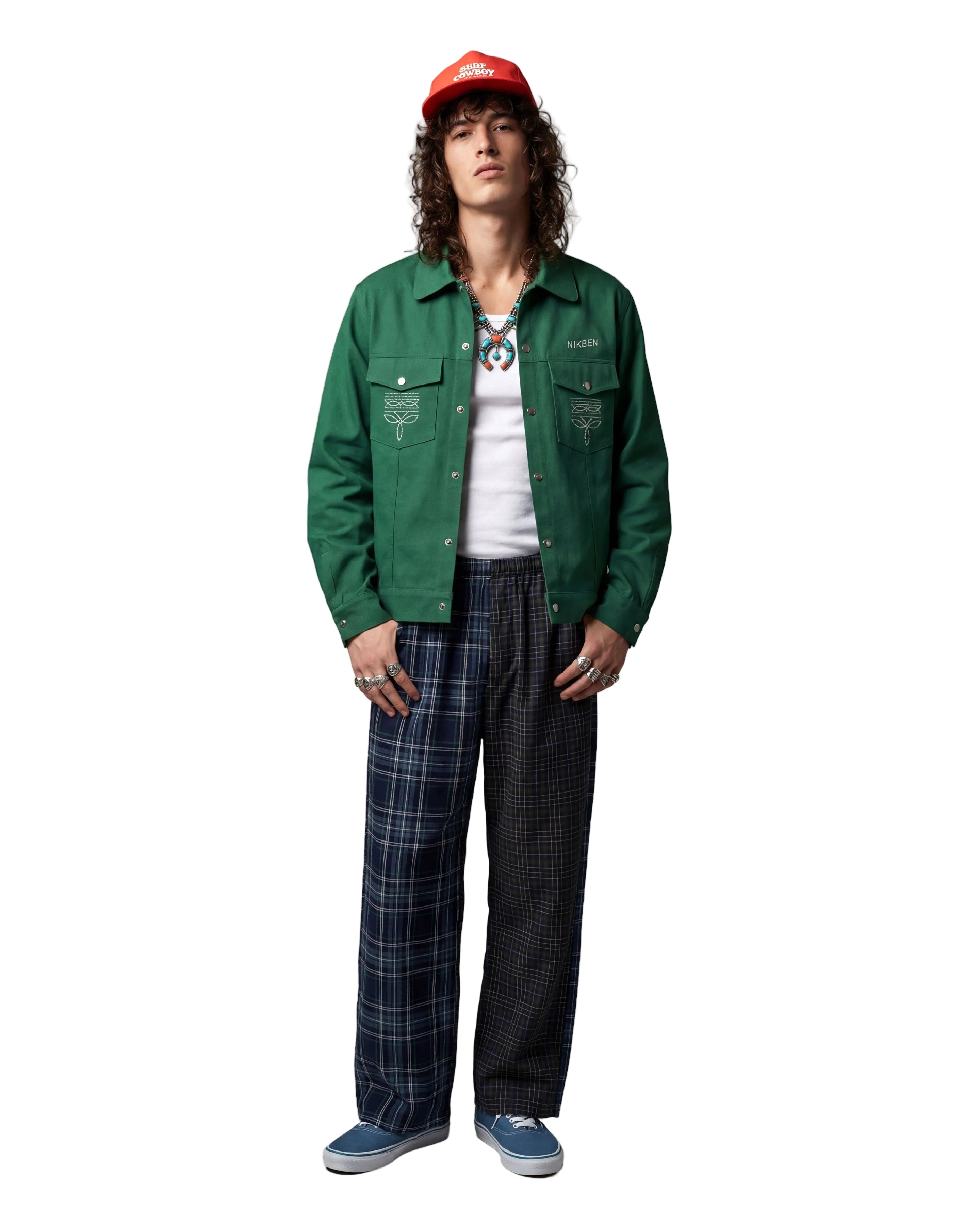 A model stands against a light gray studio background wearing a green button-up jacket with embroidered chest details, a white t-shirt, a red cap, a geometric necklace, and plaid pants.
