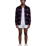A model stands against a light gray studio background wearing a dark purple and black plaid button-up shirt over a white t-shirt, white shorts, and pink bunny slippers.