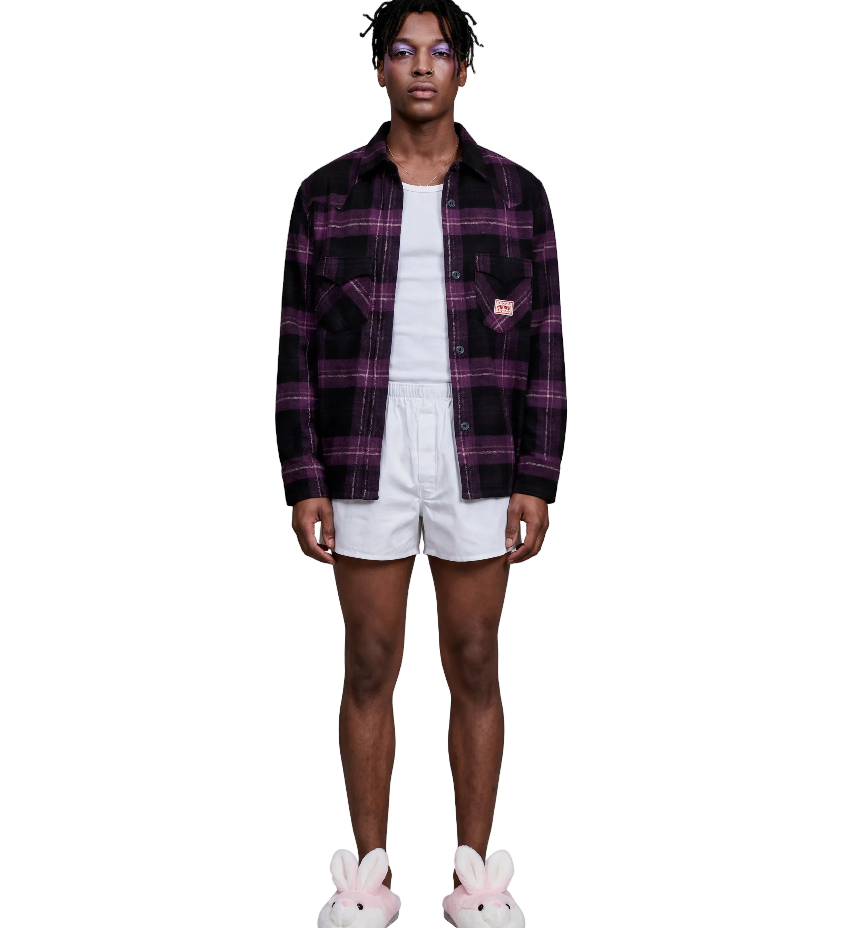 A model stands against a light gray studio background wearing a dark purple and black plaid button-up shirt over a white t-shirt, white shorts, and pink bunny slippers.