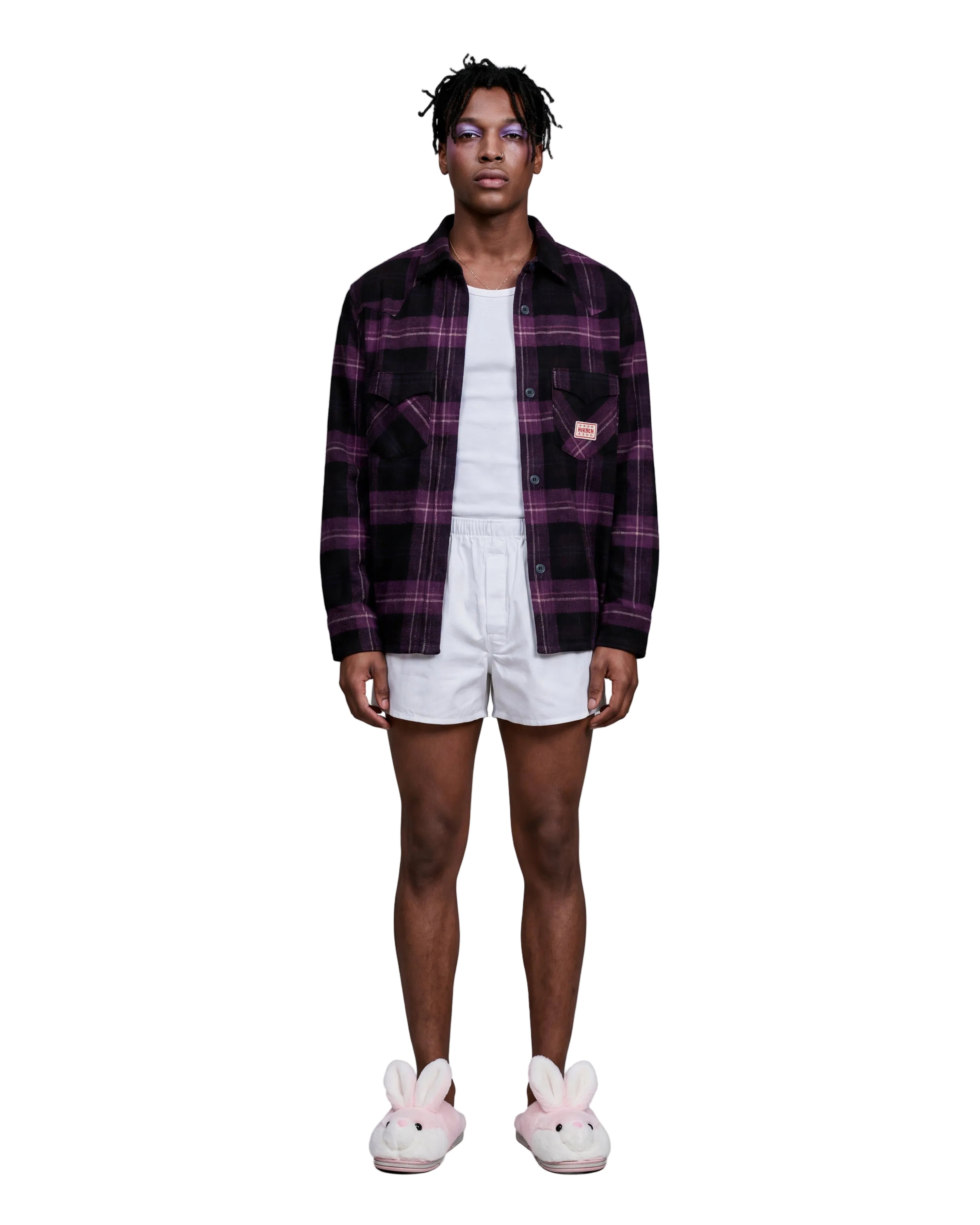 A model stands against a light gray studio background wearing a dark purple and black plaid button-up shirt over a white t-shirt, white shorts, and pink bunny slippers.