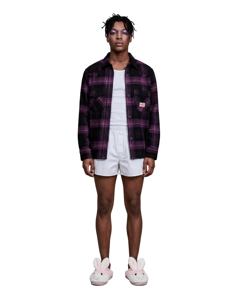 A model stands against a light gray studio background wearing a dark purple and black plaid button-up shirt over a white t-shirt, white shorts, and pink bunny slippers.