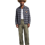 A man modeling a long-sleeve plaid button-up shirt in blue, gray, and black, worn open over a white t-shirt, paired with olive green pants and a tan cowboy hat, against a light gray background.