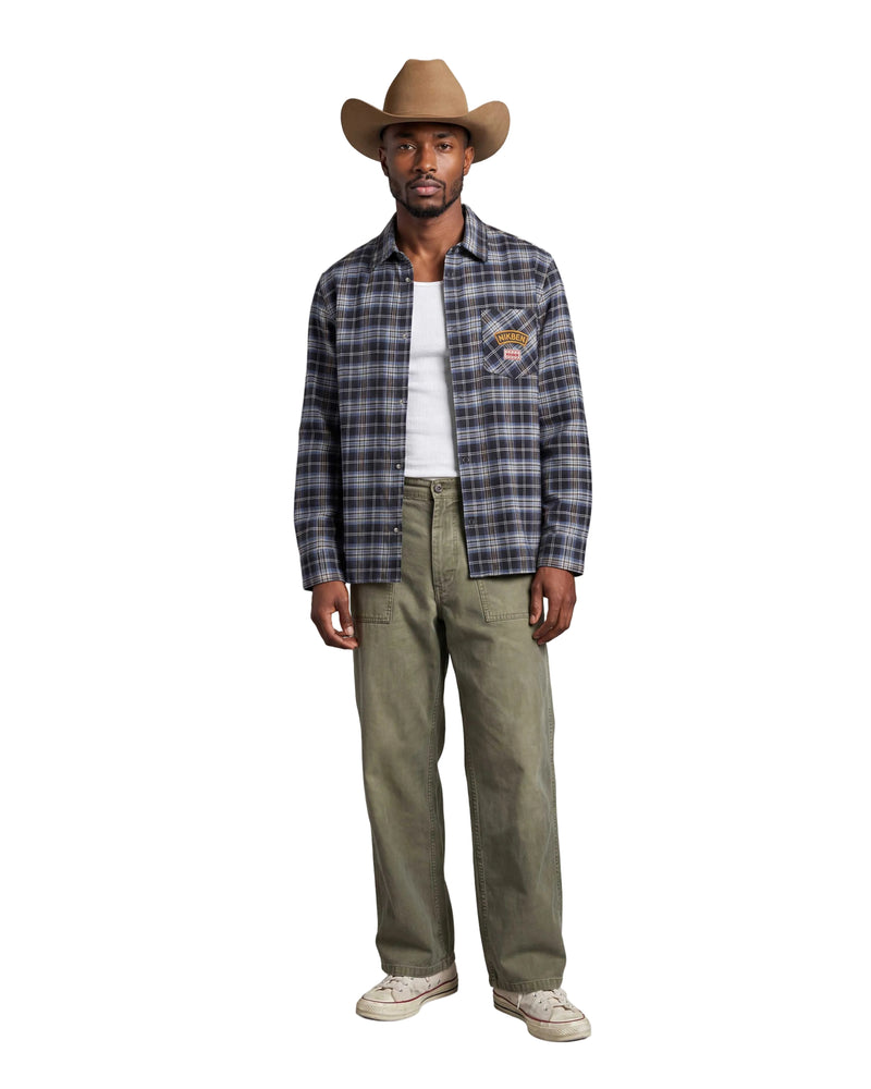 A man modeling a long-sleeve plaid button-up shirt in blue, gray, and black, worn open over a white t-shirt, paired with olive green pants and a tan cowboy hat, against a light gray background.