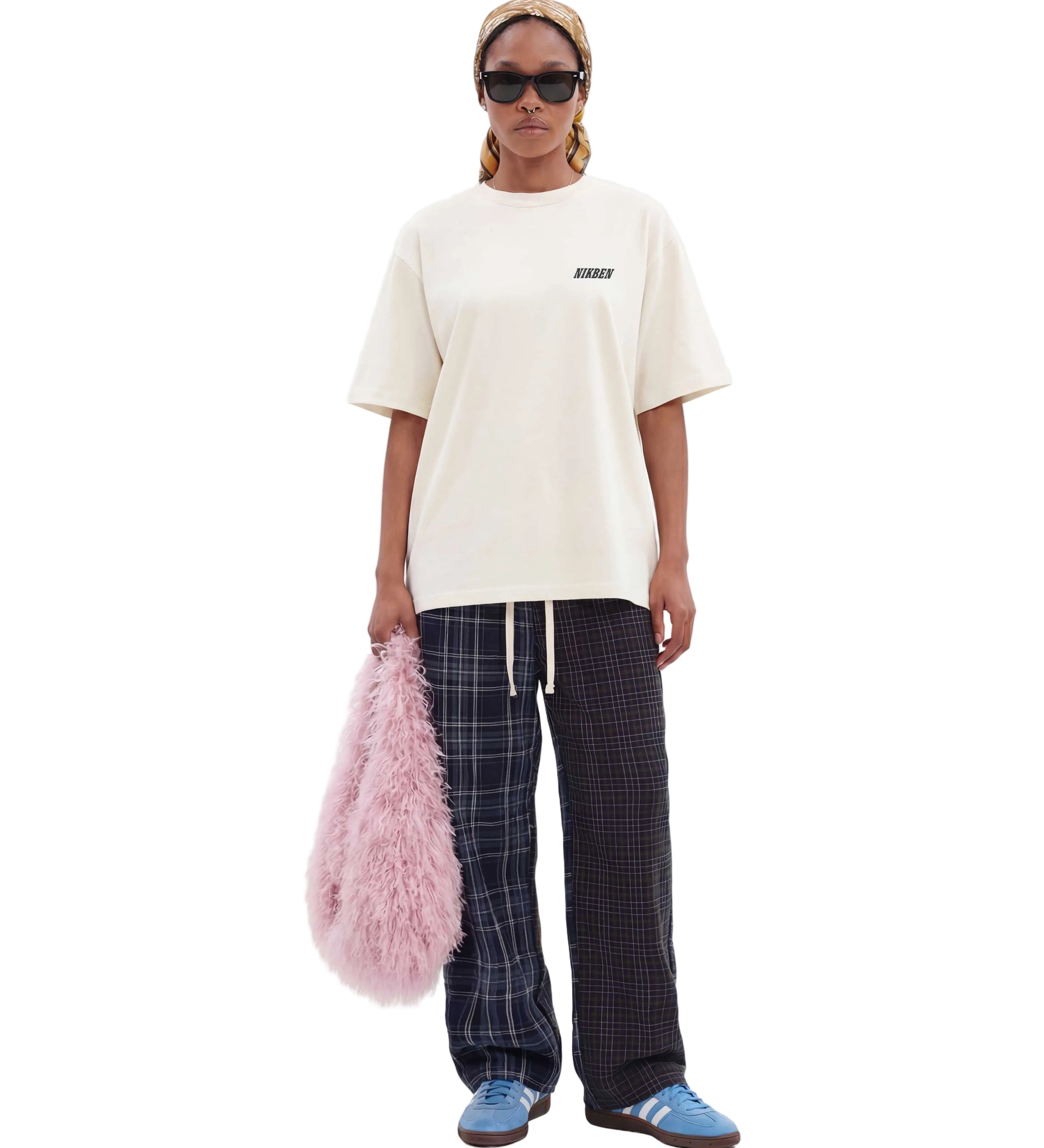 A model stands against a light gray studio background wearing an oversized cream t-shirt with a small black logo on the chest, dark plaid pants, blue sneakers, and holding a fluffy pink bag.