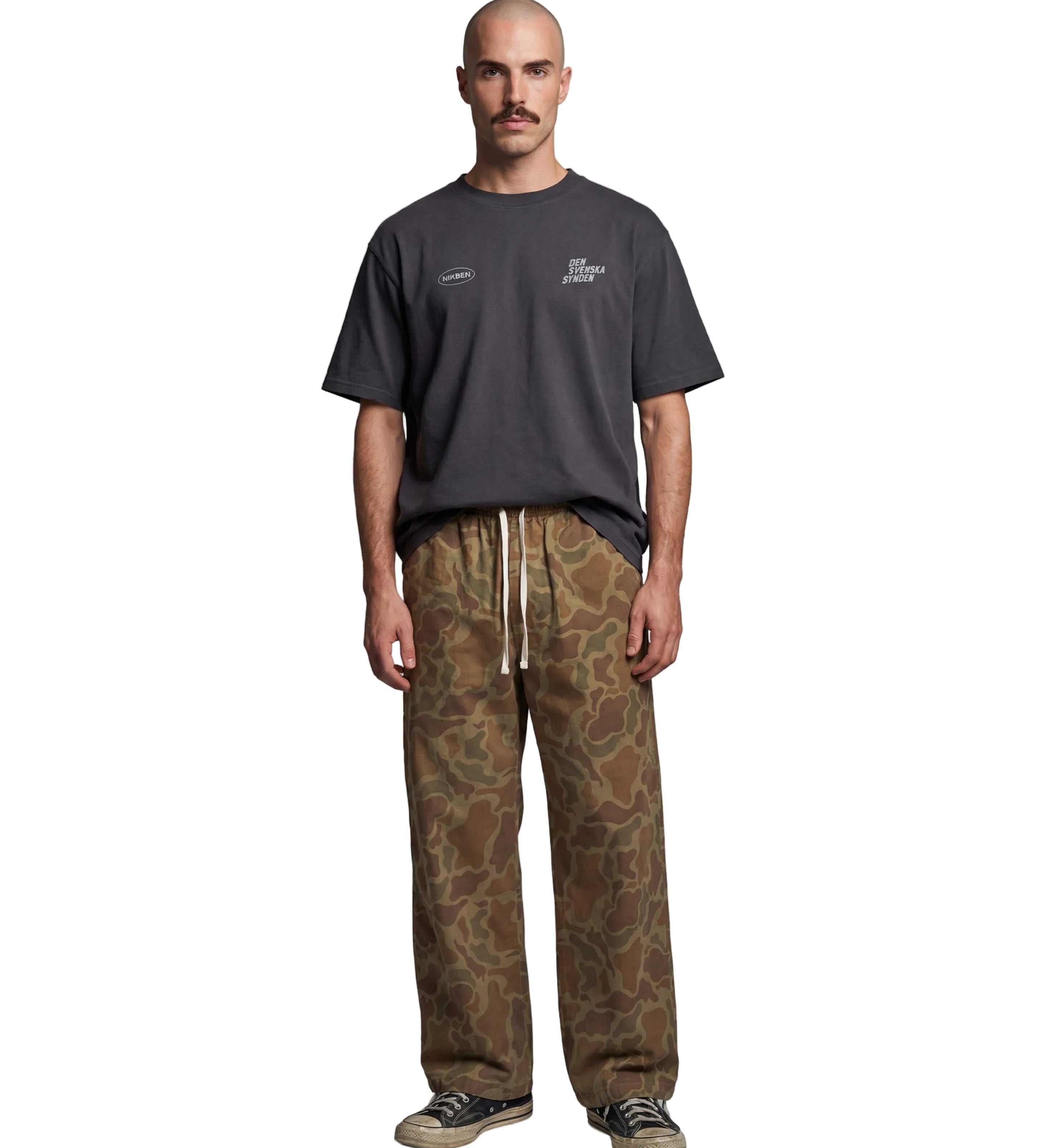 A man stands against a light gray studio background wearing a dark charcoal gray t-shirt with white printed graphics on the chest, paired with brown and beige camouflage drawstring sweatpants and black sneakers.