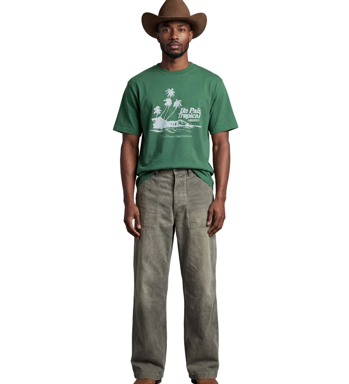 A man stands against a light gray studio background wearing a green graphic T-shirt, loose gray pants, and a brown cowboy hat. The shirt has a white palm tree and beach graphic with the text 'In The Beaches'.