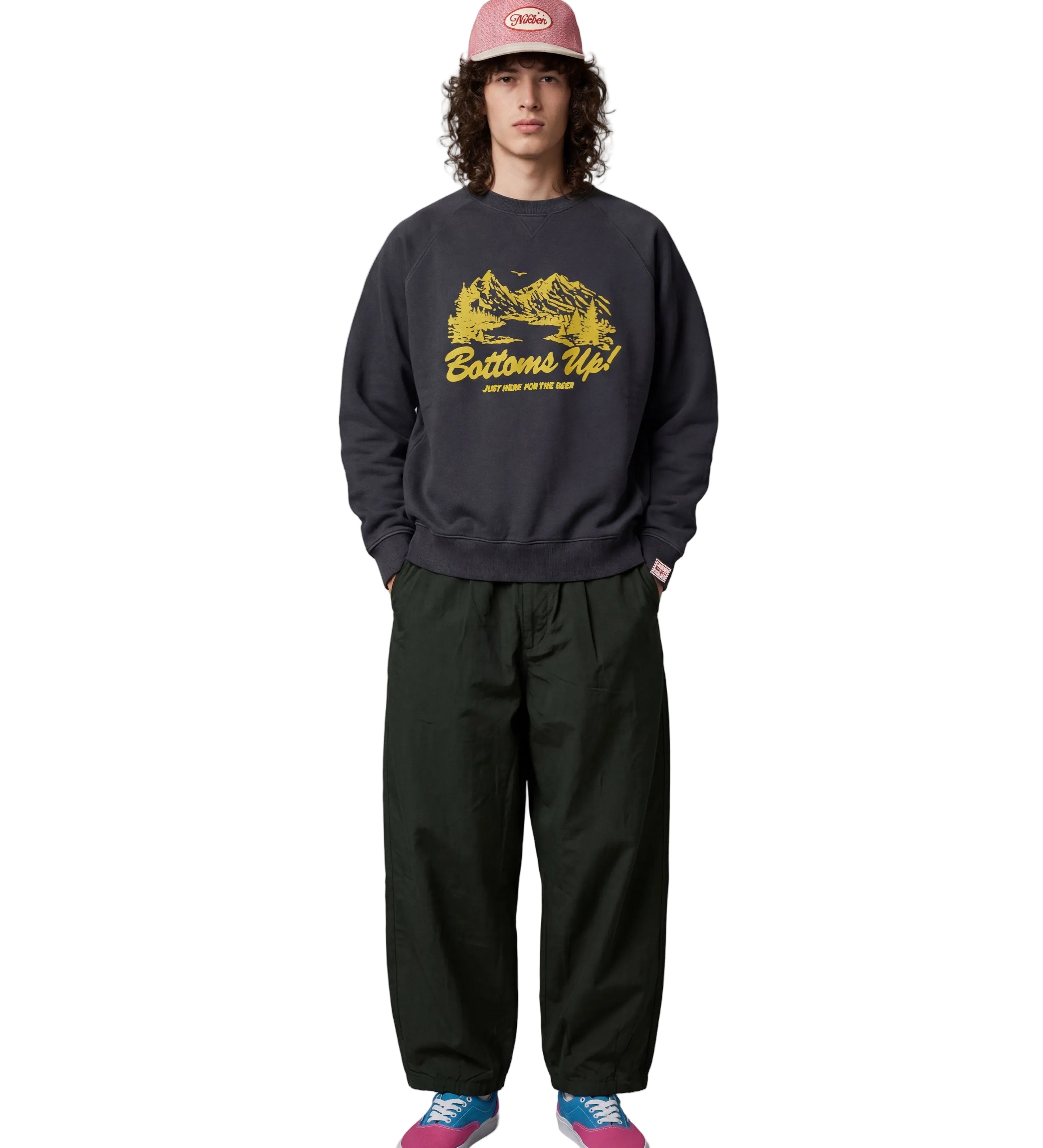 A model stands against a light gray studio background wearing a dark charcoal crewneck sweatshirt with a yellow 'Bottoms Up!' graphic, dark green baggy sweatpants, a pink cap, and pink and blue sneakers.