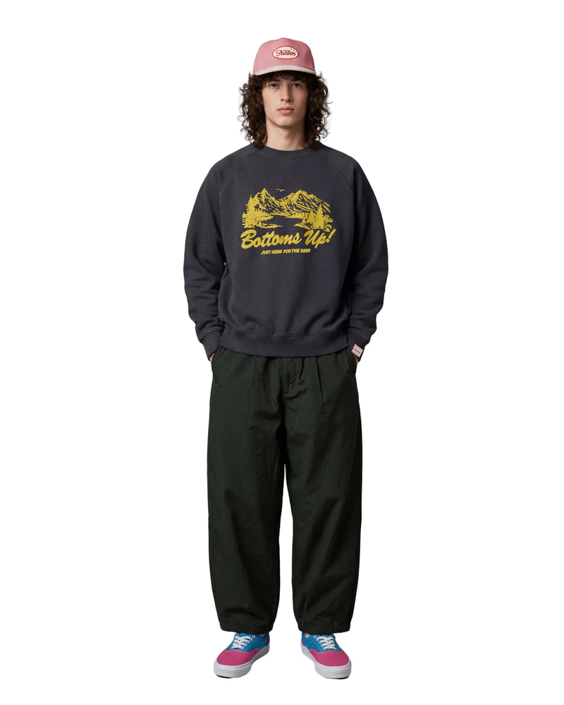 A model stands against a light gray studio background wearing a dark charcoal crewneck sweatshirt with a yellow 'Bottoms Up!' graphic, dark green baggy sweatpants, a pink cap, and pink and blue sneakers.