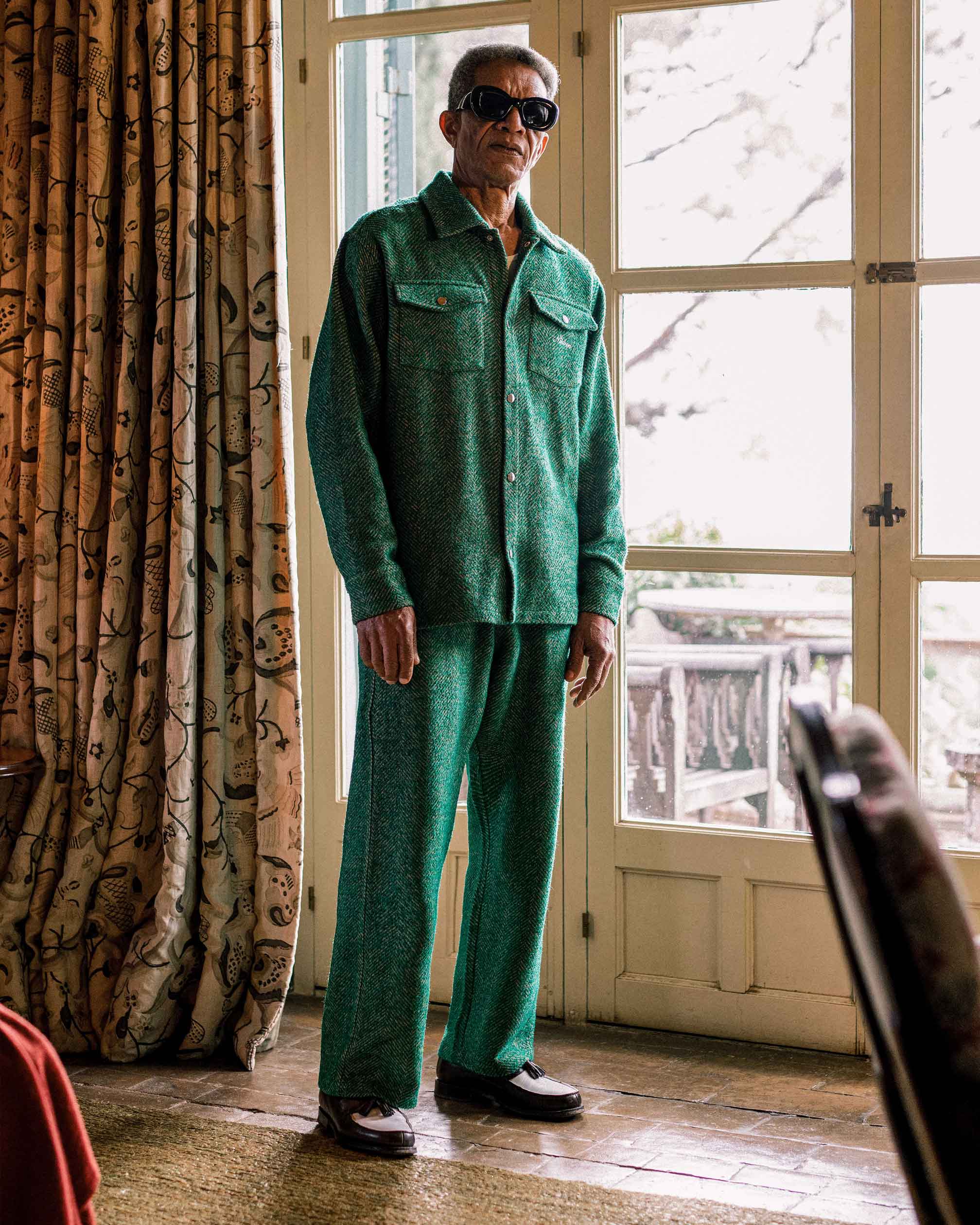Model is wearing a green outfit standing in a room with a large window and patterned curtain.
