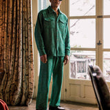Model wearing a green shirt and trouser set standing in a room with curtains and a door.