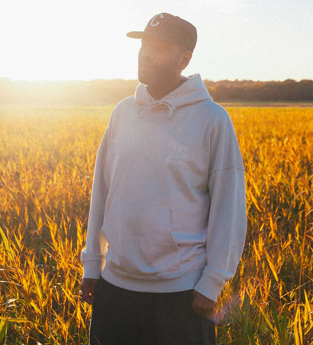 Model wearing a beige hoodie standing in a field with sunset light