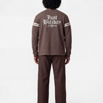 Backside of male model wearing brown longsleeve T-shirt with white "JUST HOLIDAY" print on back and white stripes on sleeves