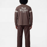 Backside of male model wearing brown longsleeve T-shirt with white "JUST HOLIDAY" print on back and white stripes on sleeves