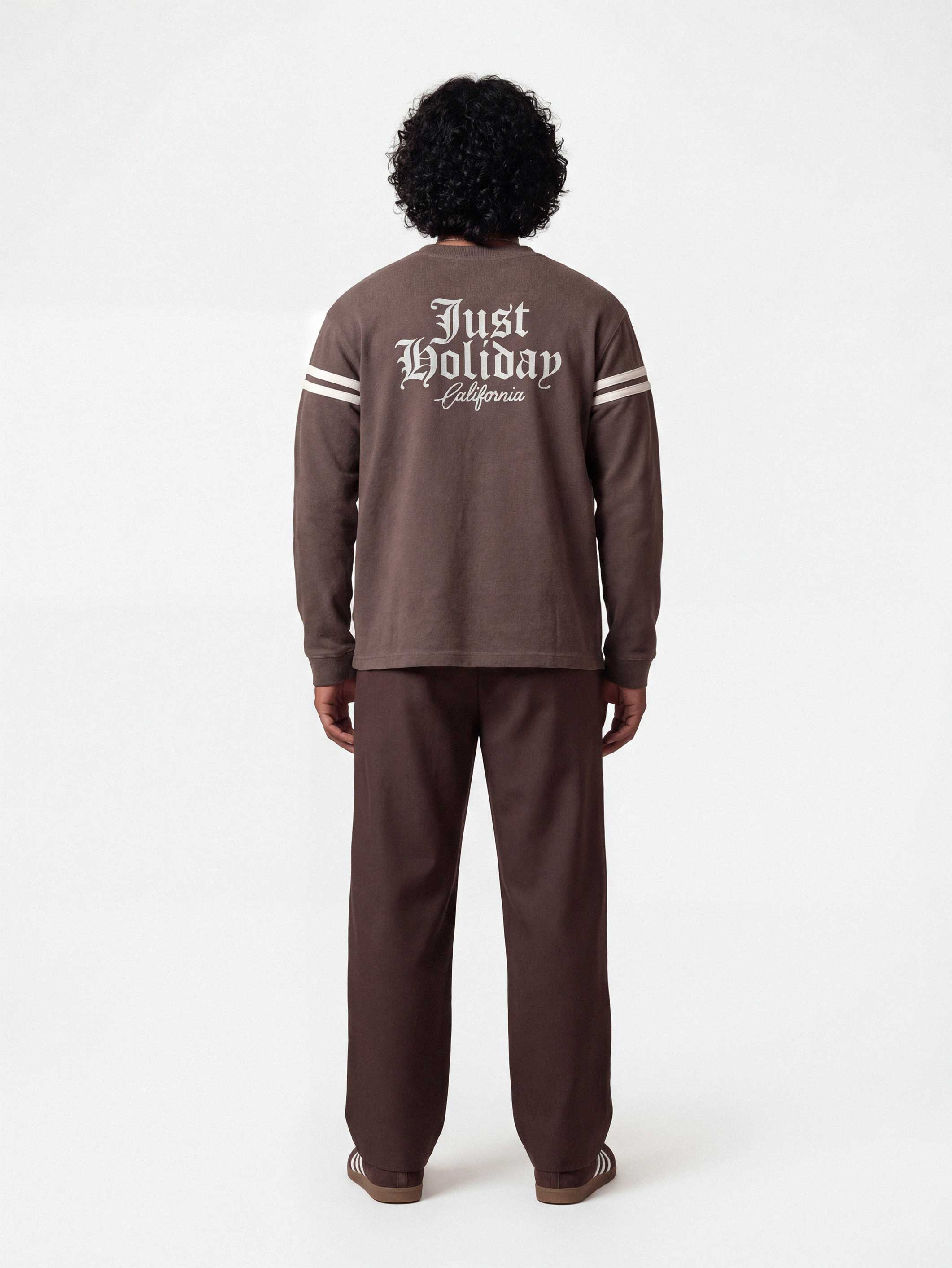 Backside of male model wearing brown longsleeve T-shirt with white "JUST HOLIDAY" print on back and white stripes on sleeves