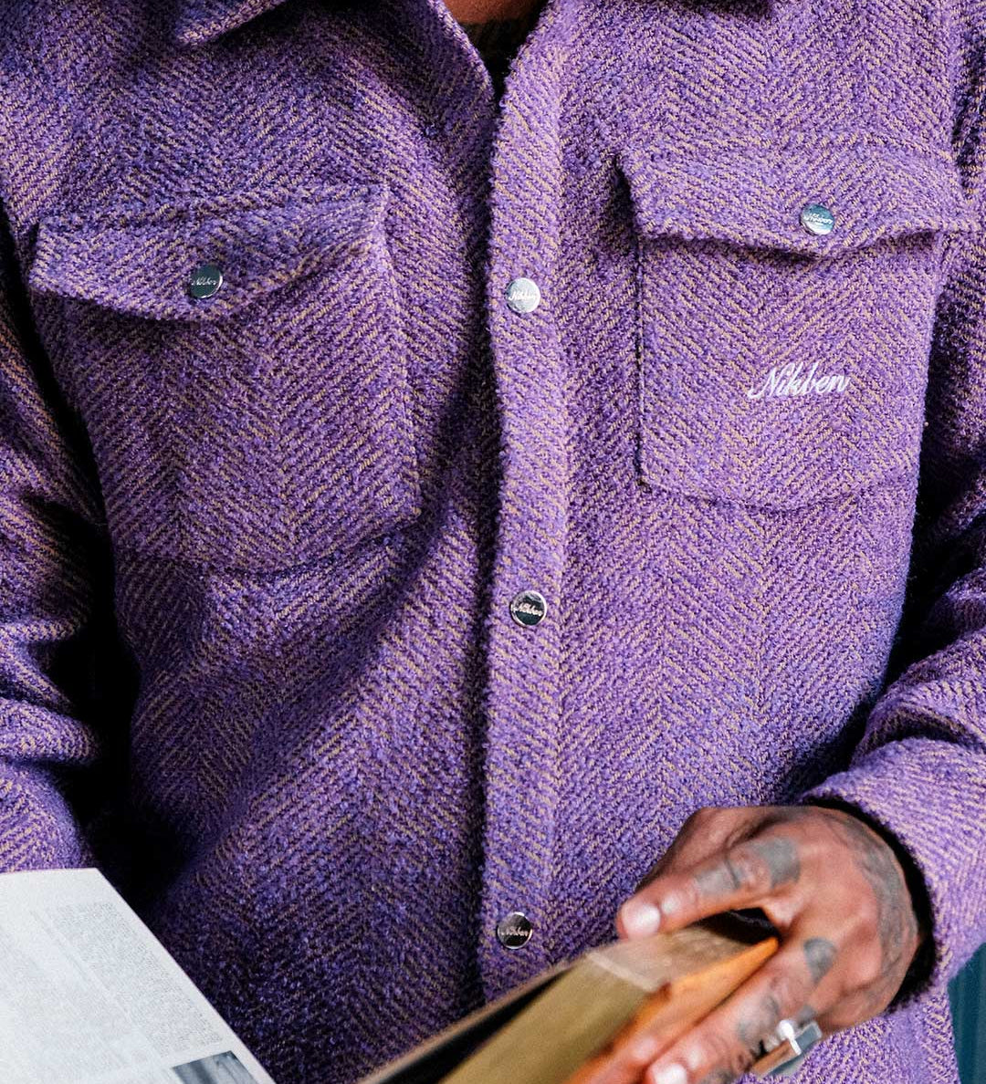 Model wearing a purple overshirt with a brand logo on pocket, holding a wooden object.