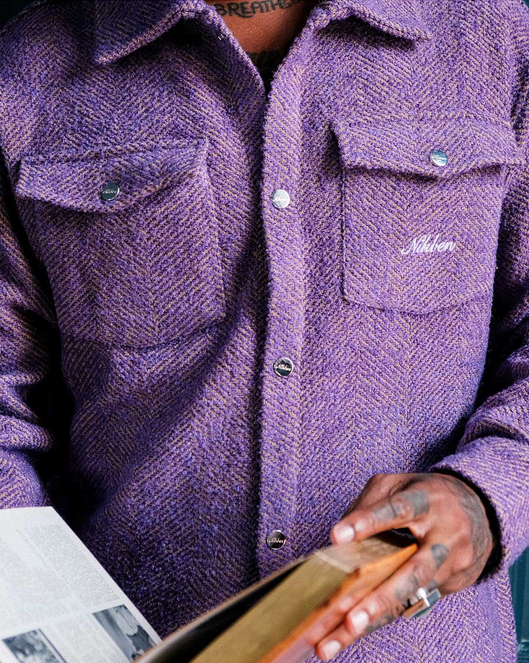 Model wearing a purple overshirt with a brand logo on pocket, holding a wooden object.