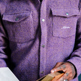 Model wearing a purple overshirt with a brand logo on pocket, holding a wooden object.