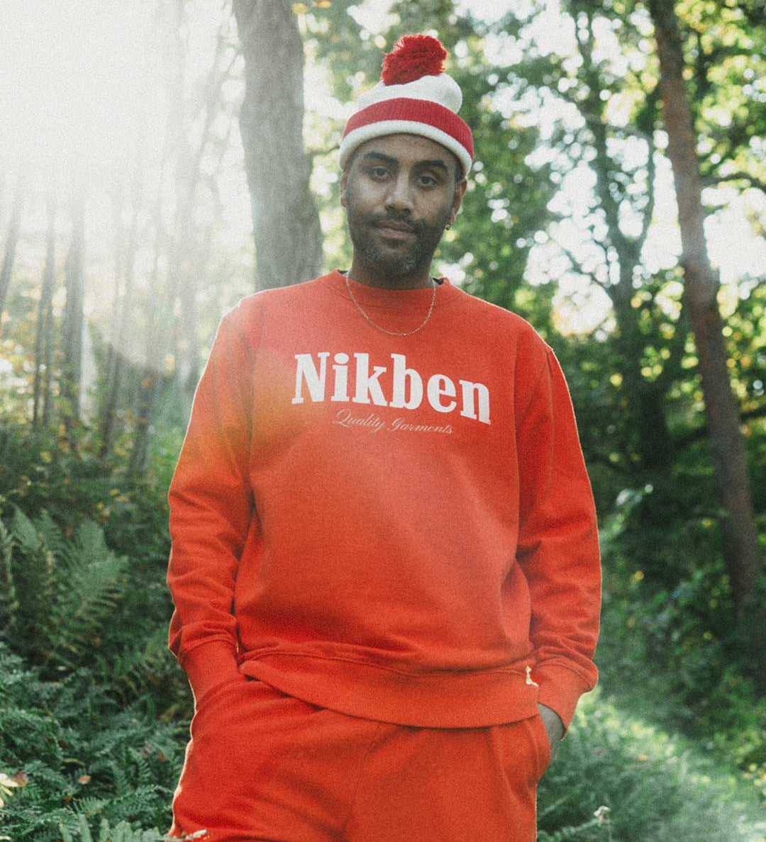 Model wearing a orange 'Nikben' sweatshirt and matching pants in a forest setting