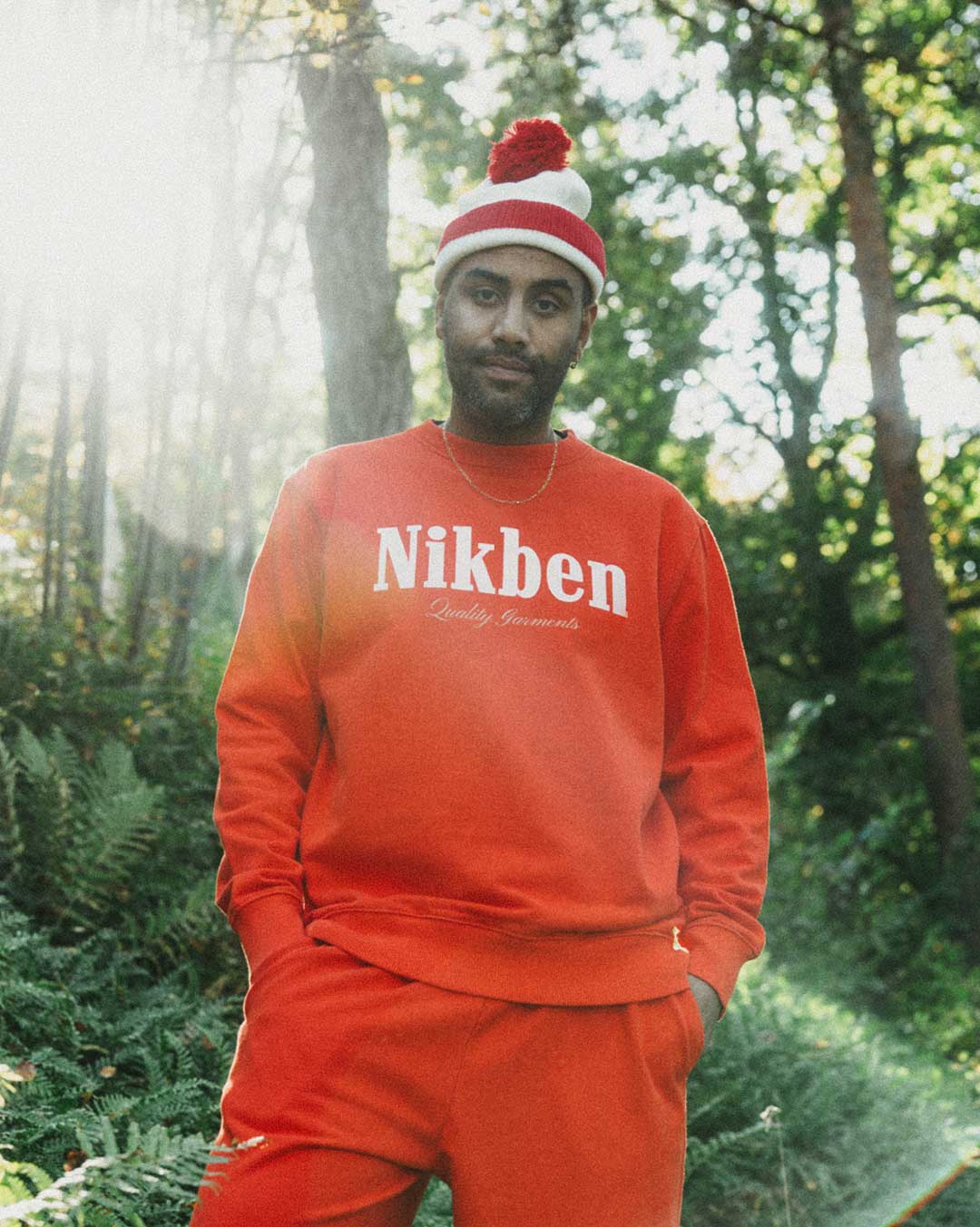 Model wearing a orange 'Nikben' sweatshirt and matching pants in a forest setting