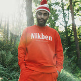 Model wearing a orange 'Nikben' sweatshirt and matching pants in a forest setting