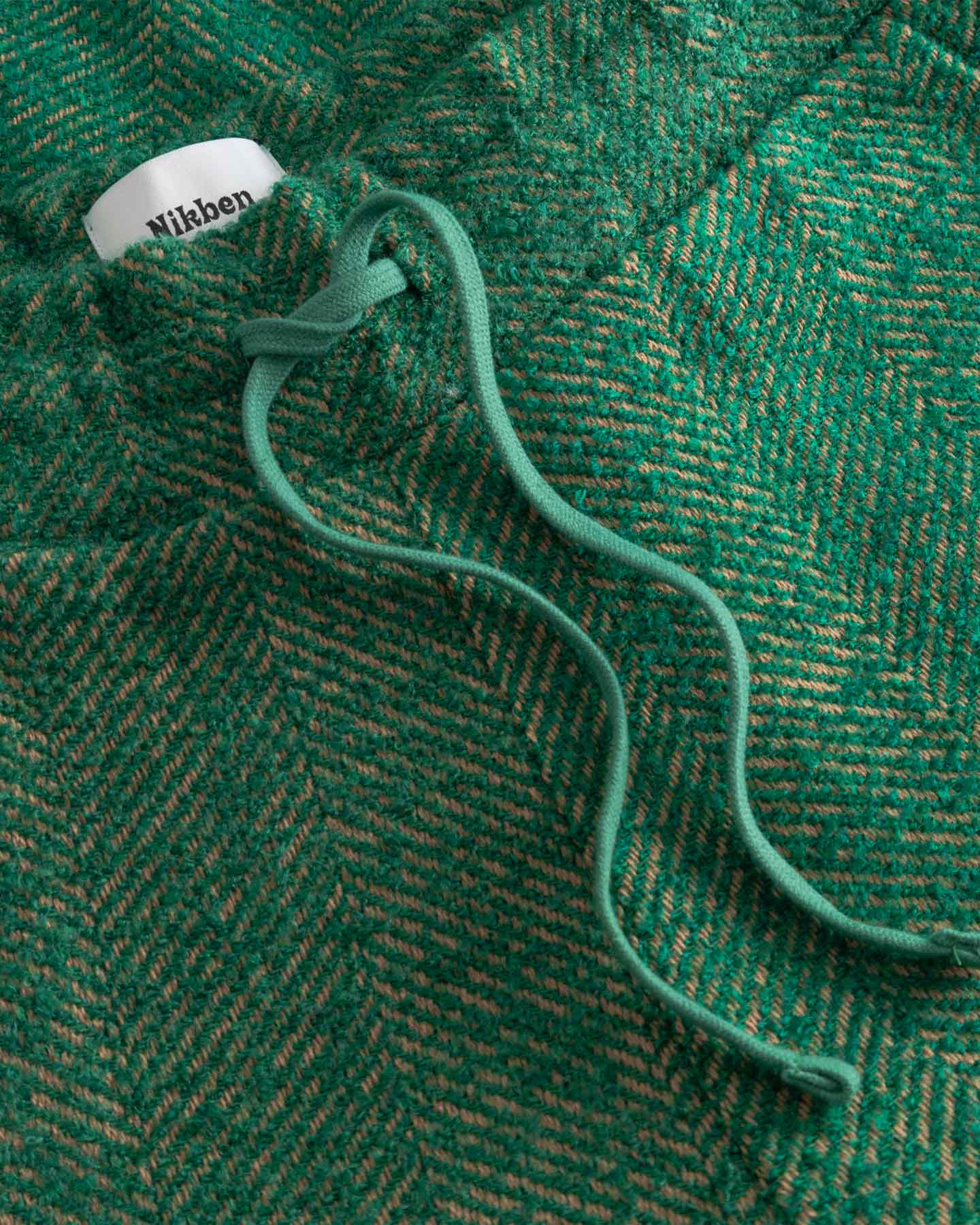 Close-up of a green textured fabric with a brand label, featuring a herringbone pattern.