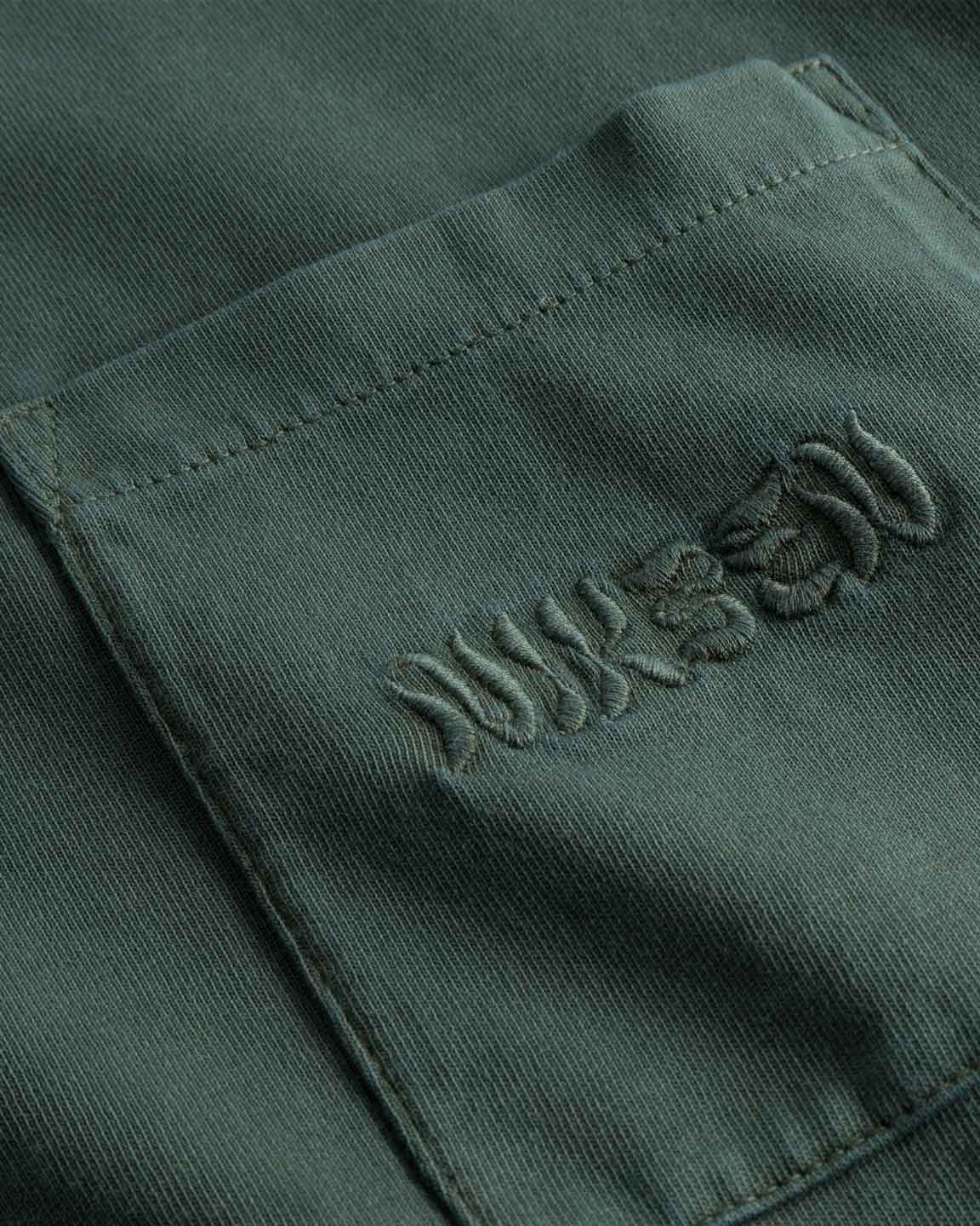 Close up of chest pocket with tone in tone embroidered logo on washed green t-shirt.