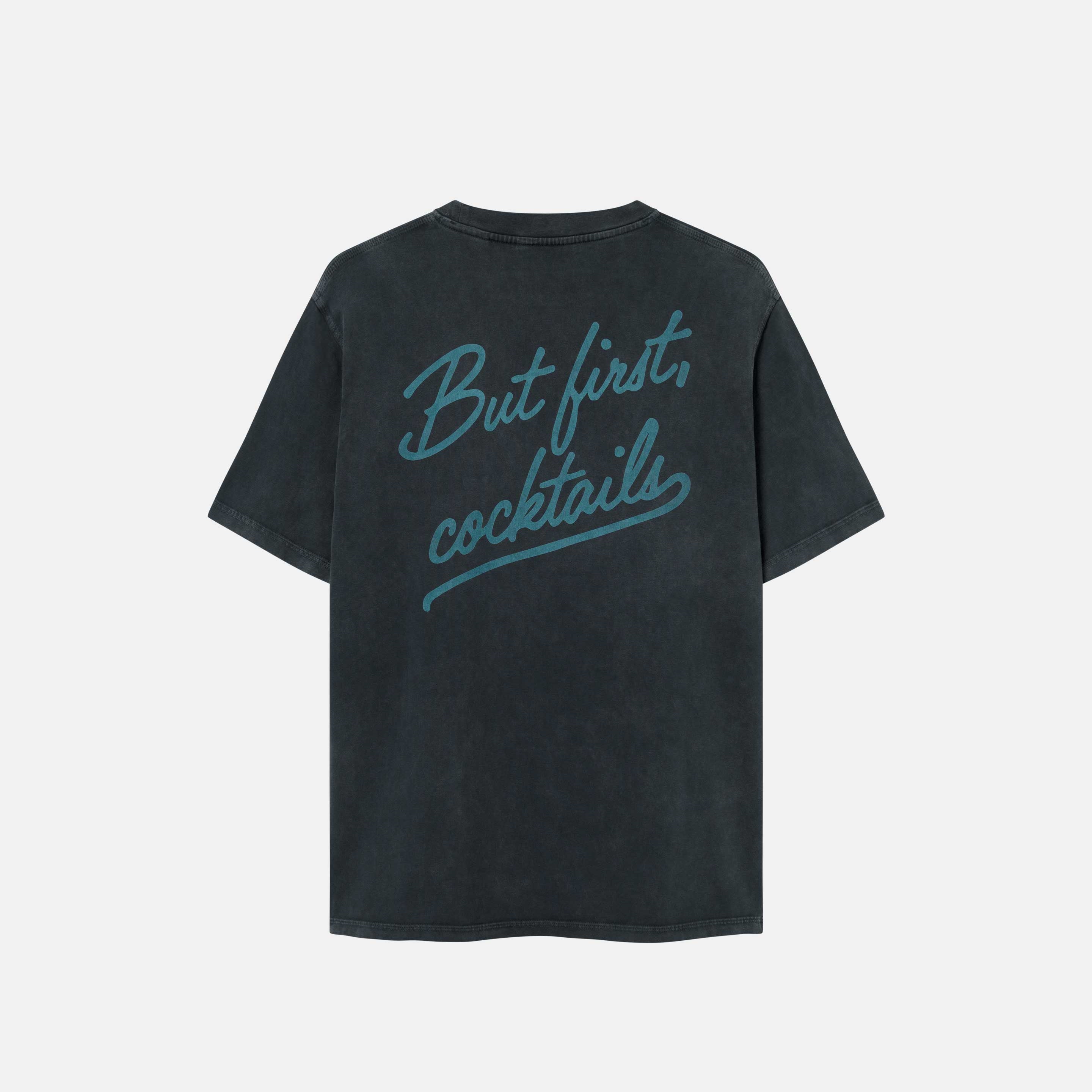 A black garment dyed t-shirt with a green "But first cocktails" text print on the back