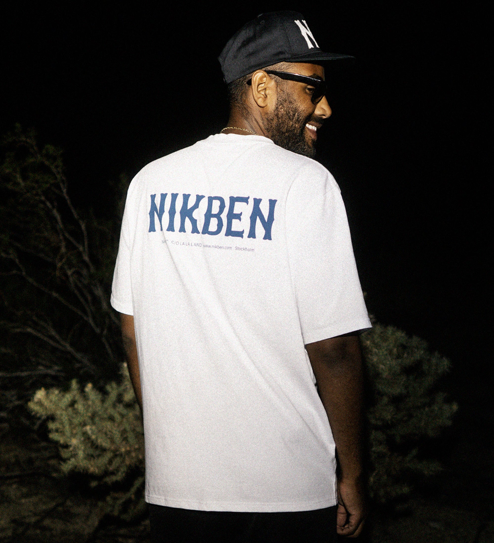Back view of male model wearing a white t-shirt with blue "Nikben" text.