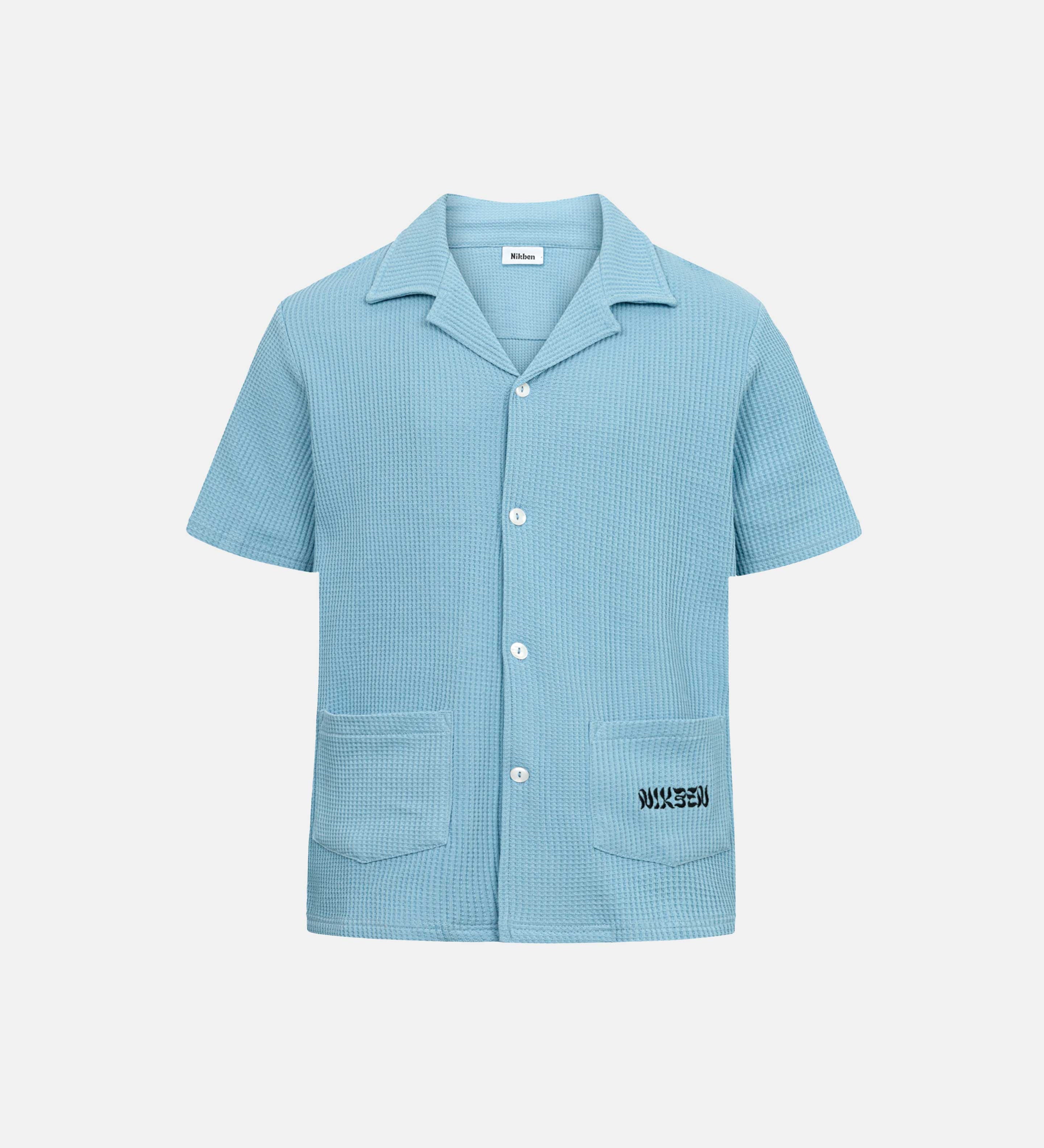 Sky blue waffle-patterned short-sleeved shirt with two front pockets, featuring a stitched black Nikben logo on the left pocket and button closure.