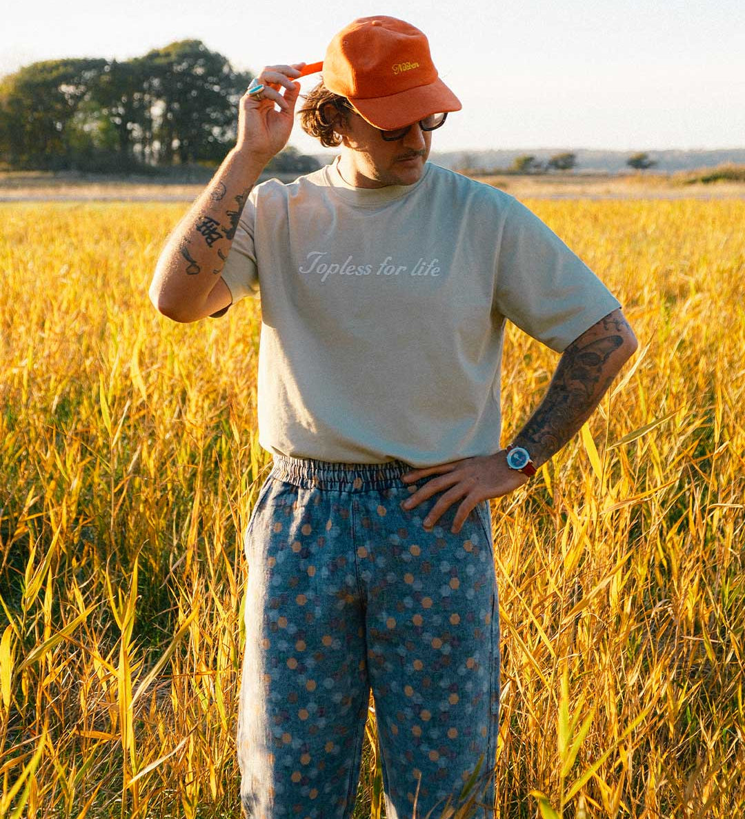 Model standing in a field wearing beige T-shirt  with text on chest and orange cap 