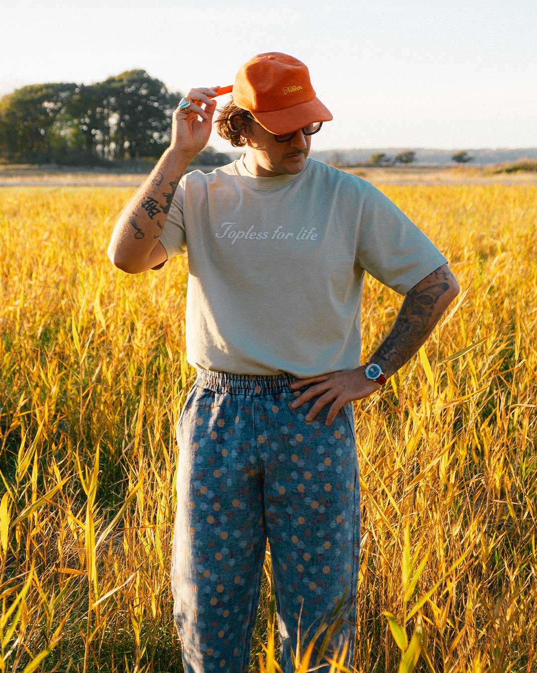 Model standing in a field wearing beige T-shirt  with text on chest and orange cap 