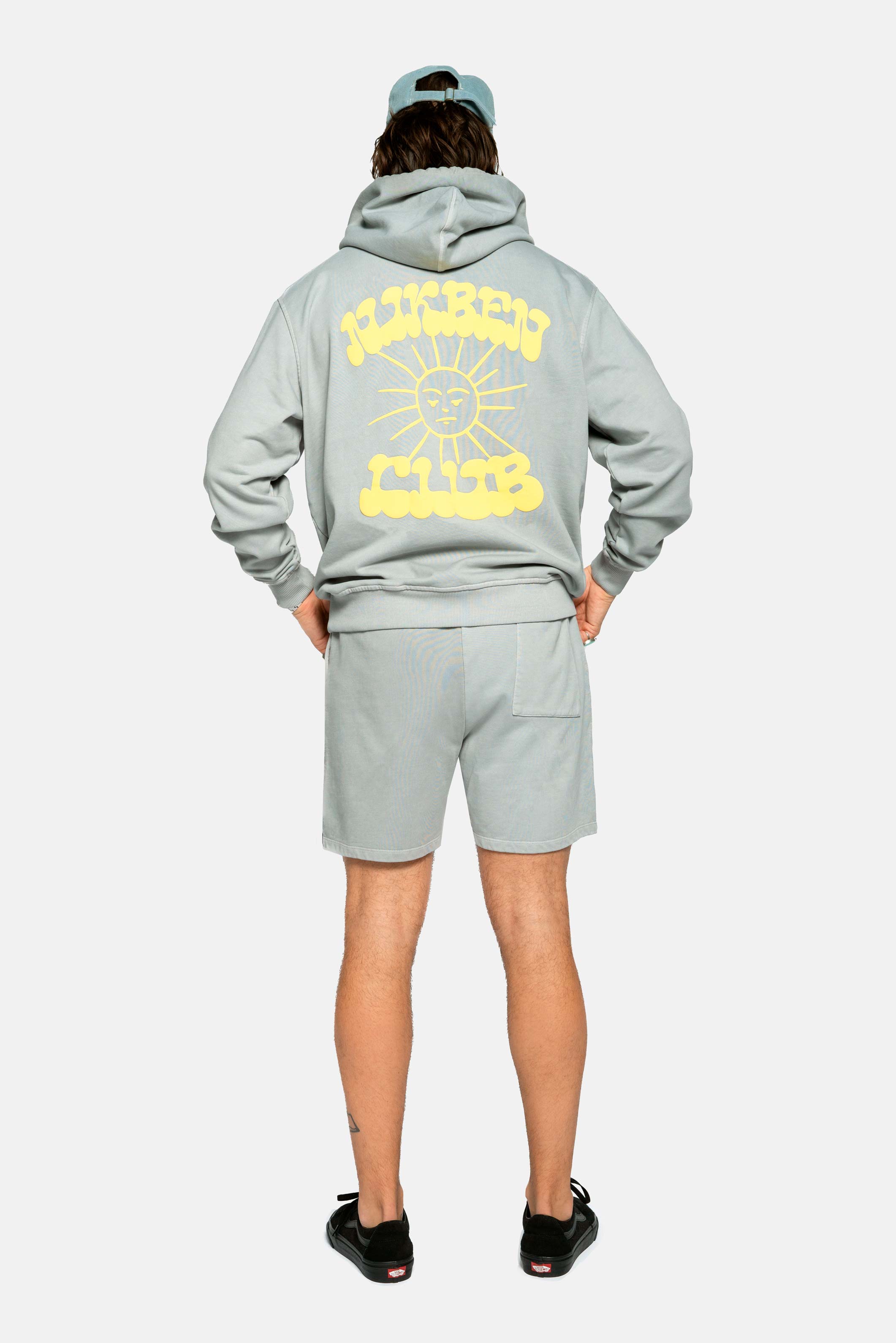 Back of Male model wearing grey hoodie with  drawstrings, chest pocket, ribbed cuffs and yellow Nikben sunshine print