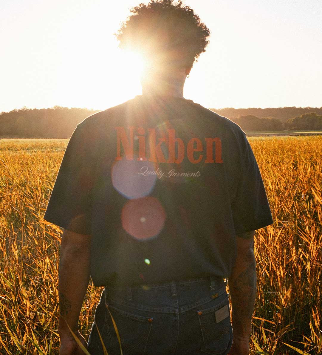 Model wearing a black t-shirt with 'Nikben' logo standing in a field at sunset.