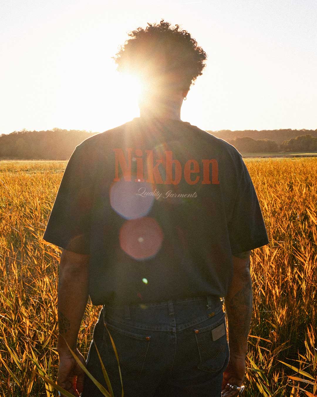 Model wearing a black t-shirt with 'Nikben' logo standing in a field at sunset.