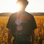 Model wearing a black t-shirt with 'Nikben' logo standing in a field at sunset.